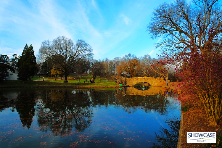 Myers Park Charlotte NC A serene scene at Myers Park in Charlotte, North Carolina, showcasing a historic stone bridge over a calm, reflective pond surrounded by blooming trees and a landscaped park. Visitors enjoy a peaceful day under a bright blue sky.