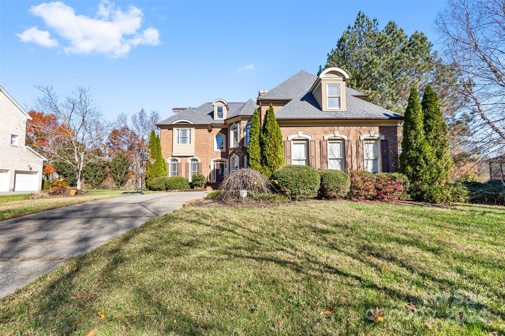The beautiful front exterior of a luxury home in Huntersville, NC at 320 Three Greens Dr, featuring a stately brick facade, elegant dormer windows, manicured landscaping, a curved driveway, and a well-maintained lawn with mature trees...