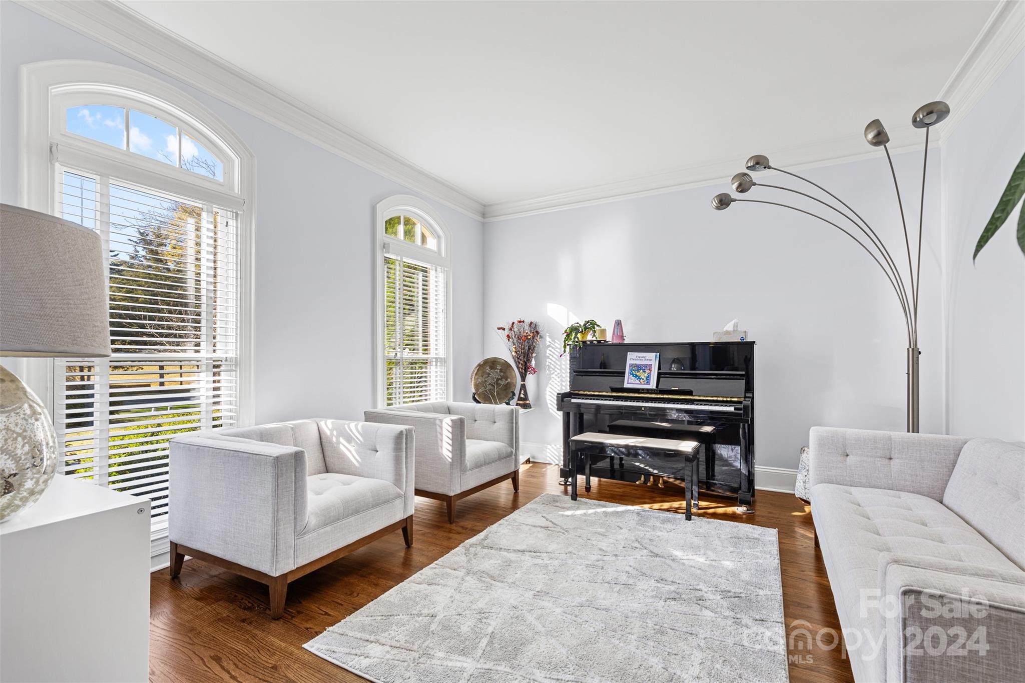 A bright and elegant sitting room in a Huntersville, NC home for sale at 320 Three Greens Dr, featuring arched windows, white tufted chairs, a modern sofa, a grand piano, and a stylish floor lamp, all set against natural light and hardwood floors...