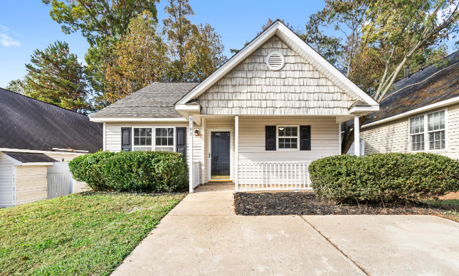 The charming front exterior of a Huntersville, NC home for sale at 320 Three Greens Dr, showcasing a single-story cottage-style home with a welcoming covered porch, well-manicured bushes, and a concrete driveway leading to the entrance...