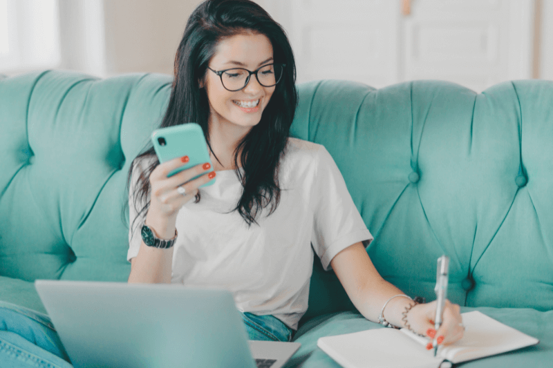 A smiling woman sitting on a teal couch using her phone and laptop while taking notes, researching Charlotte NC Homes for Sale. Represents home buyers and investors working with top real estate agents like Nancy Braun at Showcase Realty in Charlotte, North Carolina.
