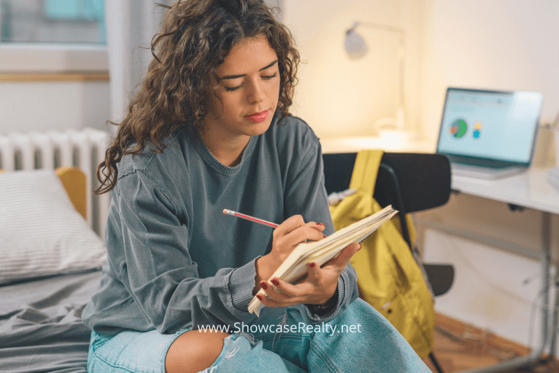 A young woman sitting on her bed, writing in a notebook with a focused expression. A laptop and workspace are visible in the background, suggesting research or planning related to home investments in Charlotte NC, assisted by Showcase Realty and top real estate agents like Nancy Braun.

