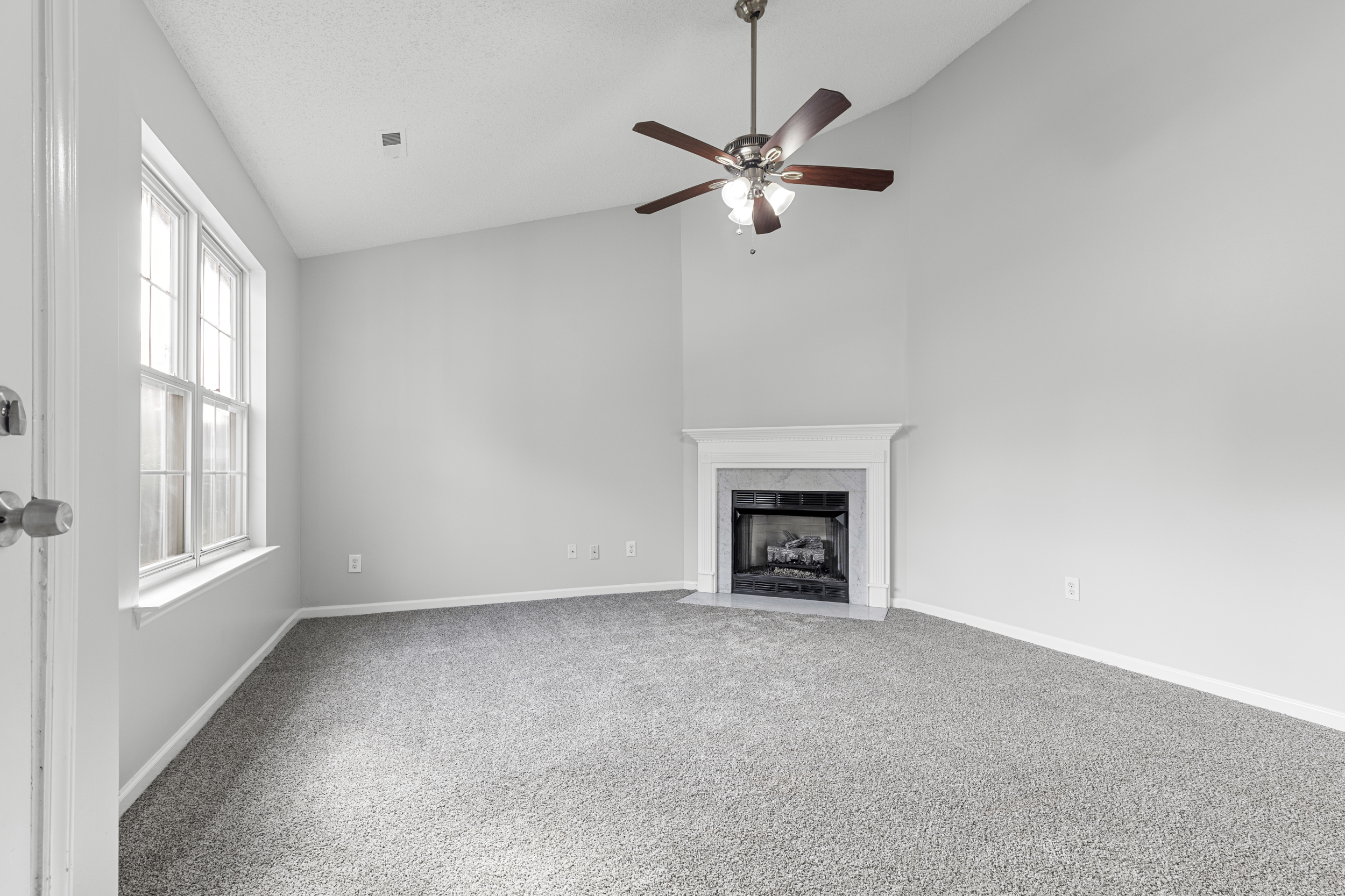 A cozy living room in a Huntersville, NC home for sale at 320 Three Greens Dr, featuring a plush carpet, high vaulted ceilings, a ceiling fan, a gas fireplace with a white mantel, and large windows allowing ample natural light...
