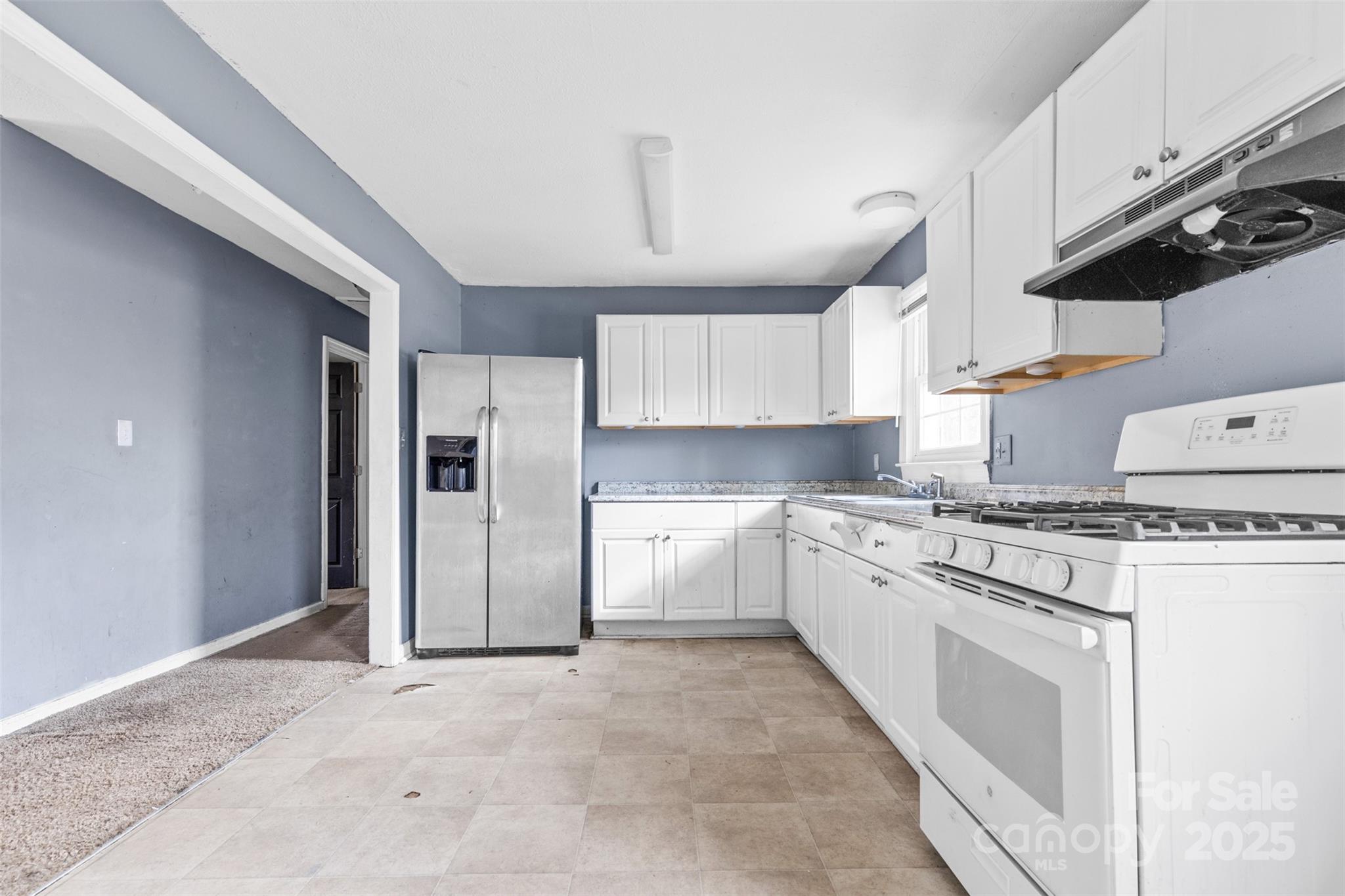 2209 Donnabrook Ln, Gastonia, NC 28052 - 3 A spacious kitchen in a Gastonia, NC home for sale at 2209 Donnabrook Ln, featuring white cabinetry, a gas stove, a stainless steel refrigerator, and blue accent walls with natural light from a nearby window...