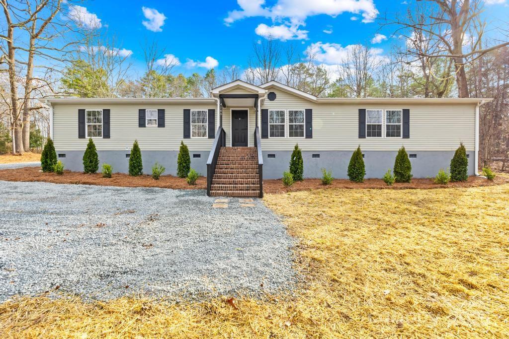 The charming front exterior of a Waxhaw, NC home for sale at 4105 Glenola Dr, showcasing a modern ranch-style home with a welcoming front porch, neutral siding, black shutters, a landscaped yard, and a gravel driveway leading up to the entrance...