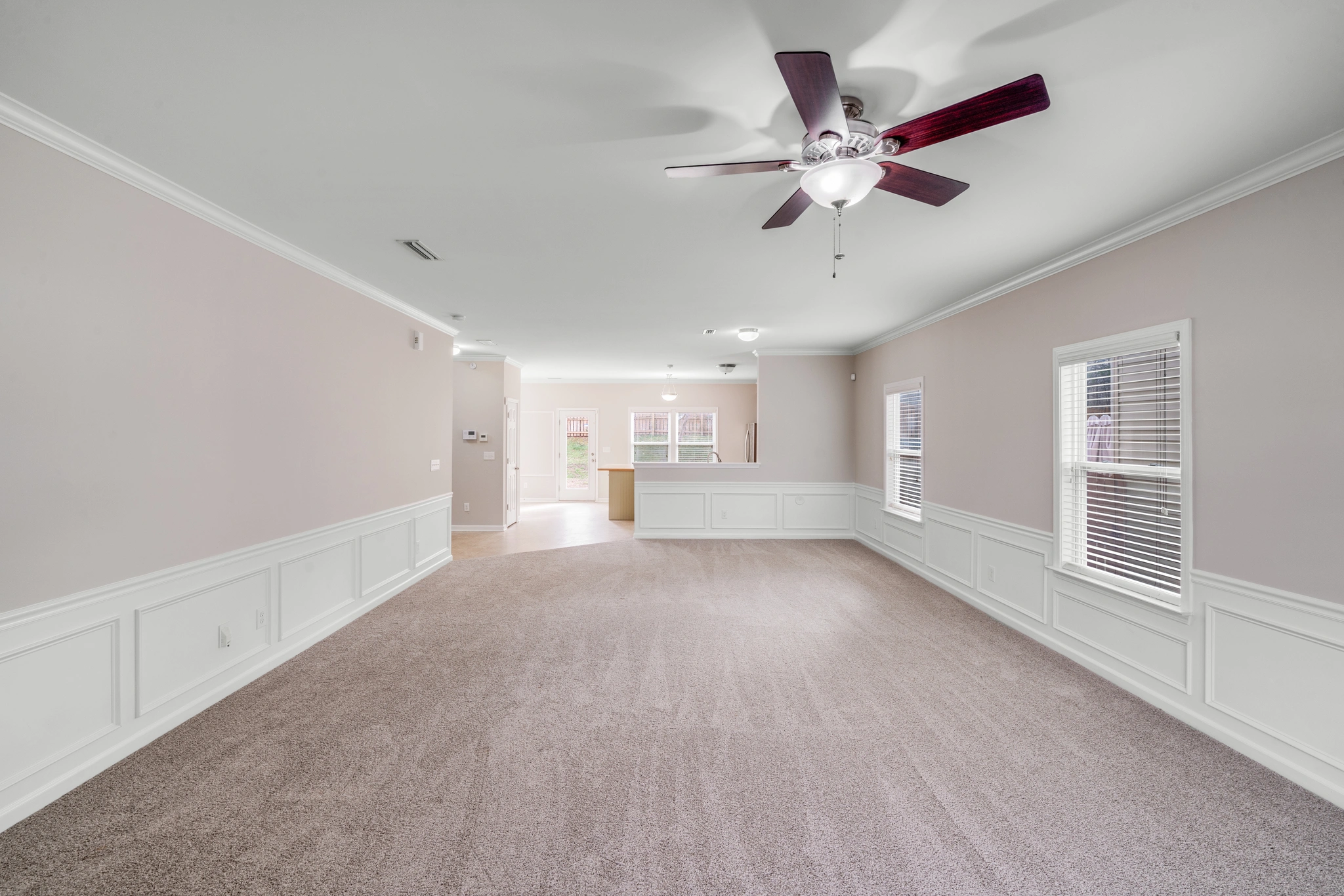 A bright and spacious living room in a Charlotte, NC home for sale at 8007 Shadbrush Ct, showcasing plush carpeting, elegant wainscoting, neutral-colored walls, large windows, and a ceiling fan for added comfort...