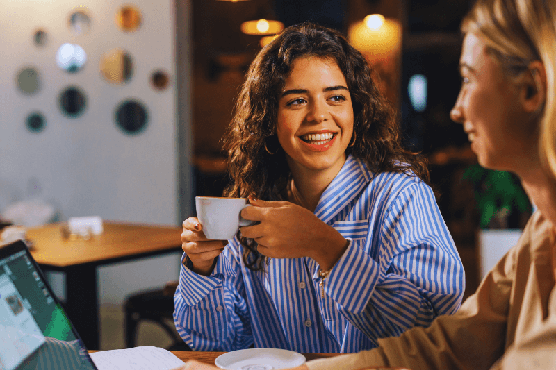 Two women sitting together with coffee, sharing a joyful conversation. This image reflects the importance of support and connection during the empty nest phase, which can be helpful when navigating Charlotte NC Real Estate decisions, especially with the expertise of Showcase Realty.
