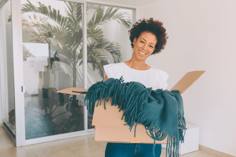 A smiling woman holding a moving box with a cozy blanket inside, standing in a bright, modern home. This image represents a new homeowner in the Charlotte housing market, reflecting the growing demand for homes in Charlotte, NC, as buyers continue to settle into their dream homes in 2025.