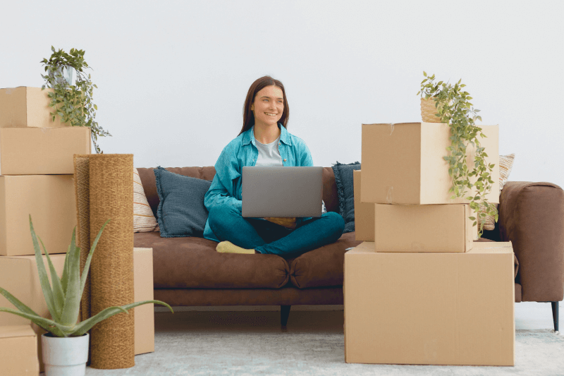 A young woman sitting on a couch with a laptop, surrounded by moving boxes and houseplants. She appears excited about settling into her new home, reflecting the strong real estate activity in the Charlotte housing market. This image highlights the trend of new homebuyers relocating to Charlotte, NC, in early 2025.