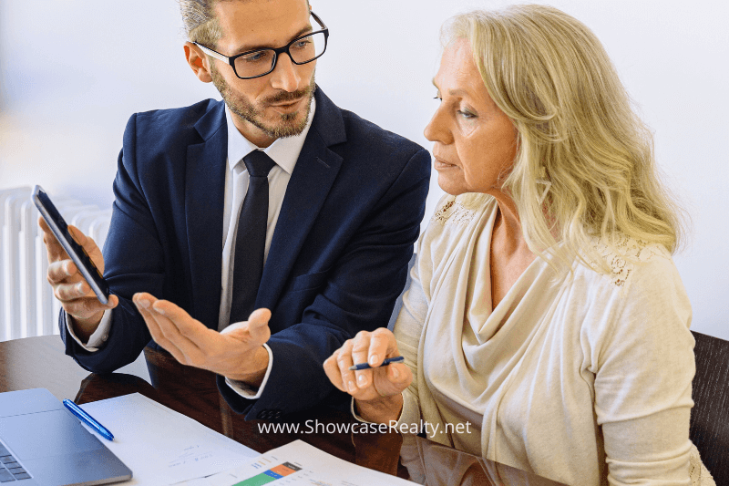 A professional-looking man in a suit is explaining financial details to an attentive older woman. He holds a smartphone while gesturing, and they are seated at a table with documents, charts, and a laptop. The woman listens closely, holding a pen, suggesting a discussion about real estate commissions or market analysis. This image reflects the expertise and personalized service provided by Charlotte NC real estate agents at Showcase Realty, highlighting financial guidance in the Charlotte real estate market.