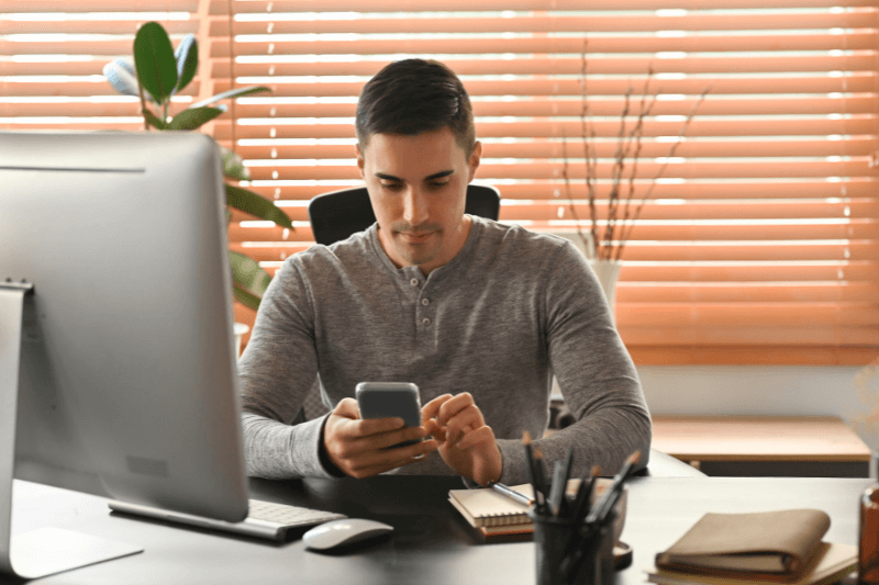 A man sitting at a modern desk using his smartphone and computer, researching Charlotte NC Real Estate and Charlotte NC Home Buying options with Showcase Realty. The image represents the home buying process, including online property searches and communication with real estate agents.