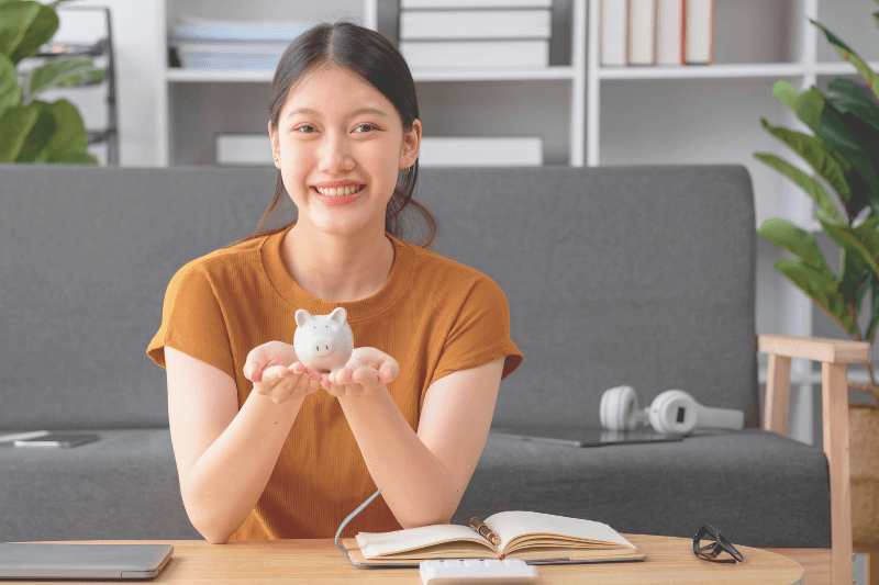 A smiling woman in a casual home setting holds a small piggy bank in her hands, symbolizing financial savings and smart investment planning. This image represents the importance of securing the right financing options for investment properties in Charlotte NC Real Estate with expert guidance from Showcase Realty.